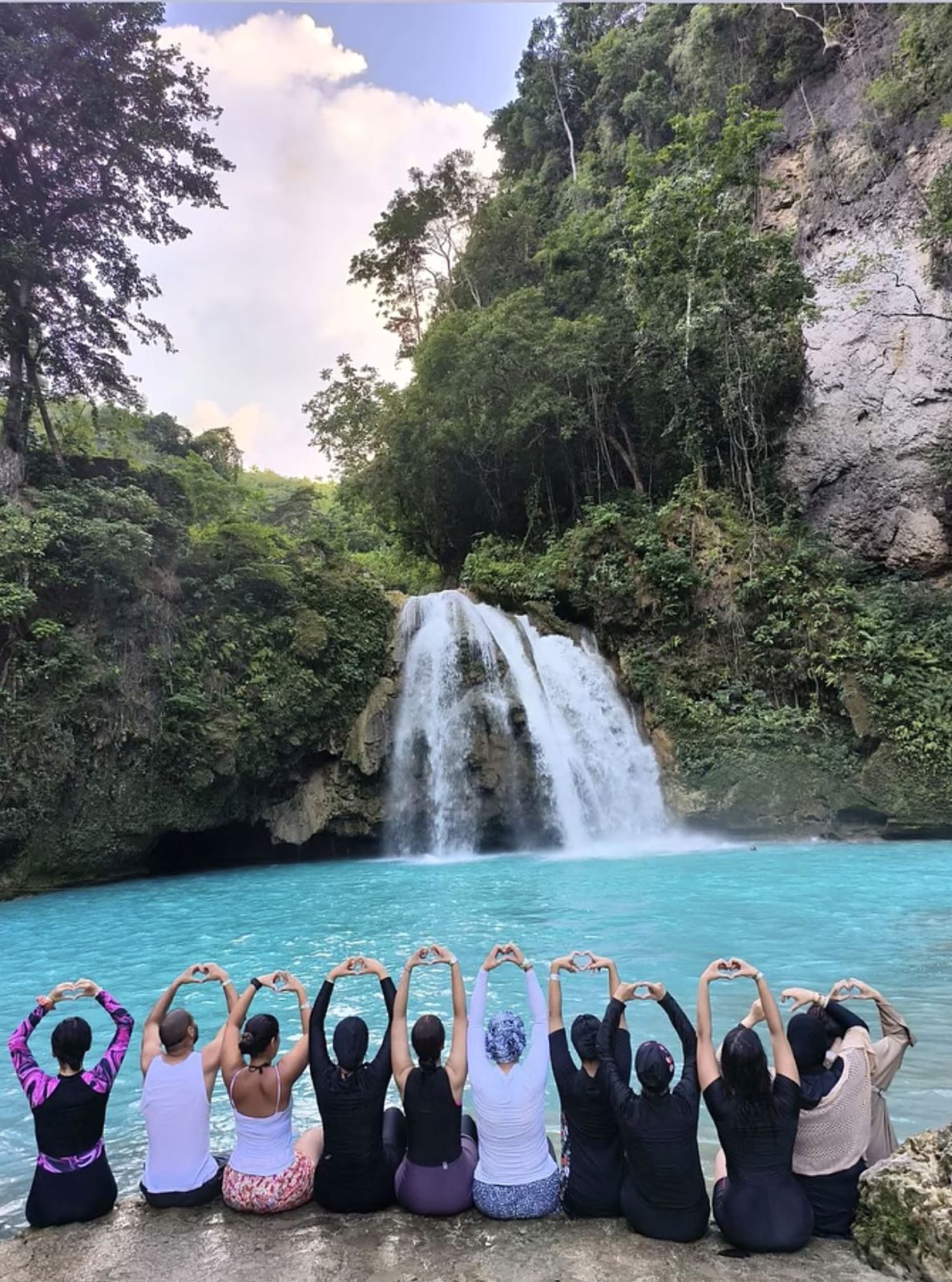 Un groupe de voyageuses Travel Horizons de dos, faisant des cœurs avec les mains devant une cascade turquoise